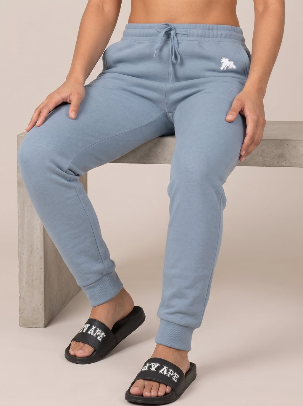 Model seated on a bench wearing black FIT APE slides and blue joggers